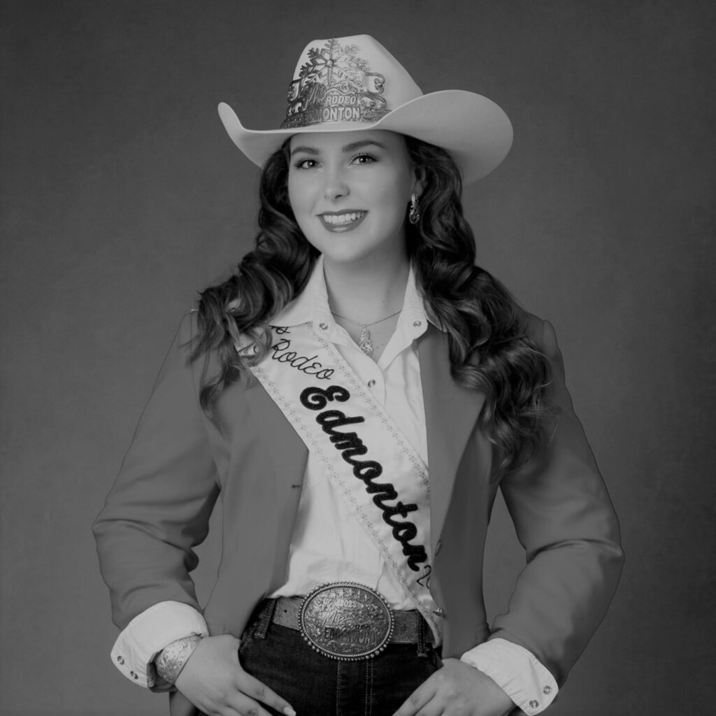 A black and white photo of Cody Wilson standing in a cowboy hat with long dark hair, wearing a blazer, a belt with a big buckle, and a sash across her chest that reads "Miss Rodeo Edmonton".